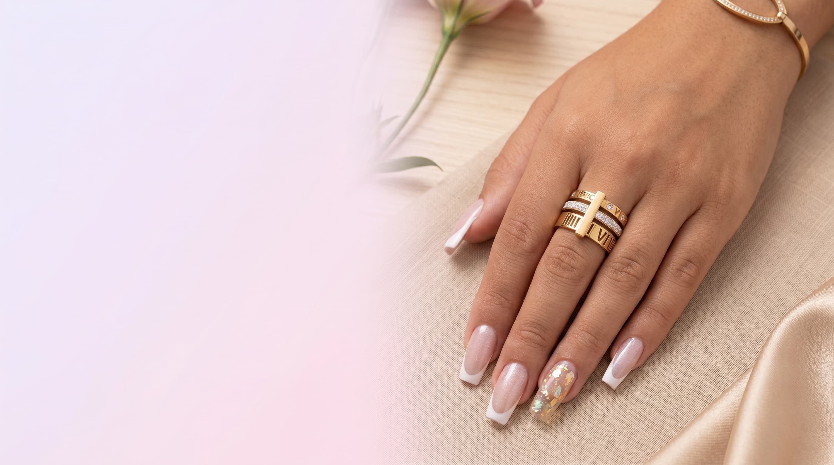 Mão feminina com unhas claras, usando um anel triplo de aço inoxidável dourado e prateado com pedras transparentes, sobre fundo desfocado.